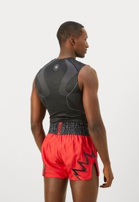 Athletic man shown from back wearing black sleeveless compression shirt and red boxing shorts with black zigzag patterns.