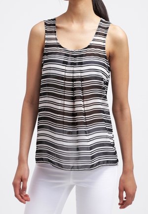 Vrouw draagt een mouwloze zwart-wit gestreepte blouse gecombineerd met een witte broek, staand tegen een egale achtergrond.