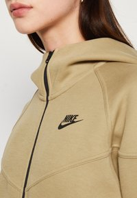 Tūlītējs Nike kapuce ar rāv�ējslēdzēju, melniem akcentiem un logo. Iezīmes mīksts, teksturēts audums un vaļīgs, mūsdienīgs dizains.
