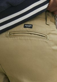 Beige Jack & Jones byxor med en bakficka, slät textur och en svart etikett med texten "JACK & JONES EST. 1990" ovanför fickan.