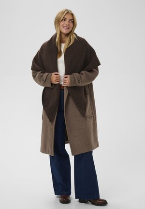 Manteau en mélange de laine brun avec un large col châle noir, grandes poches et détails en métal, associé à un jean large bleu et des bottes brunes.