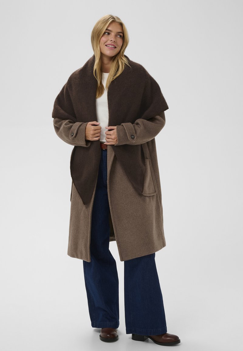 Brun uldblandingsfrakke med en bred, mørkebrun sjalkrave, store lommer og knapdetaljer, parret med blå vide jeans og brune støvler.