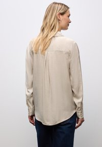 Camicia beige a maniche lunghe con una texture liscia, dotata di una piega sul retro, orlo curvo e polsini con bottoni. Abbinata a jeans blu scuro.