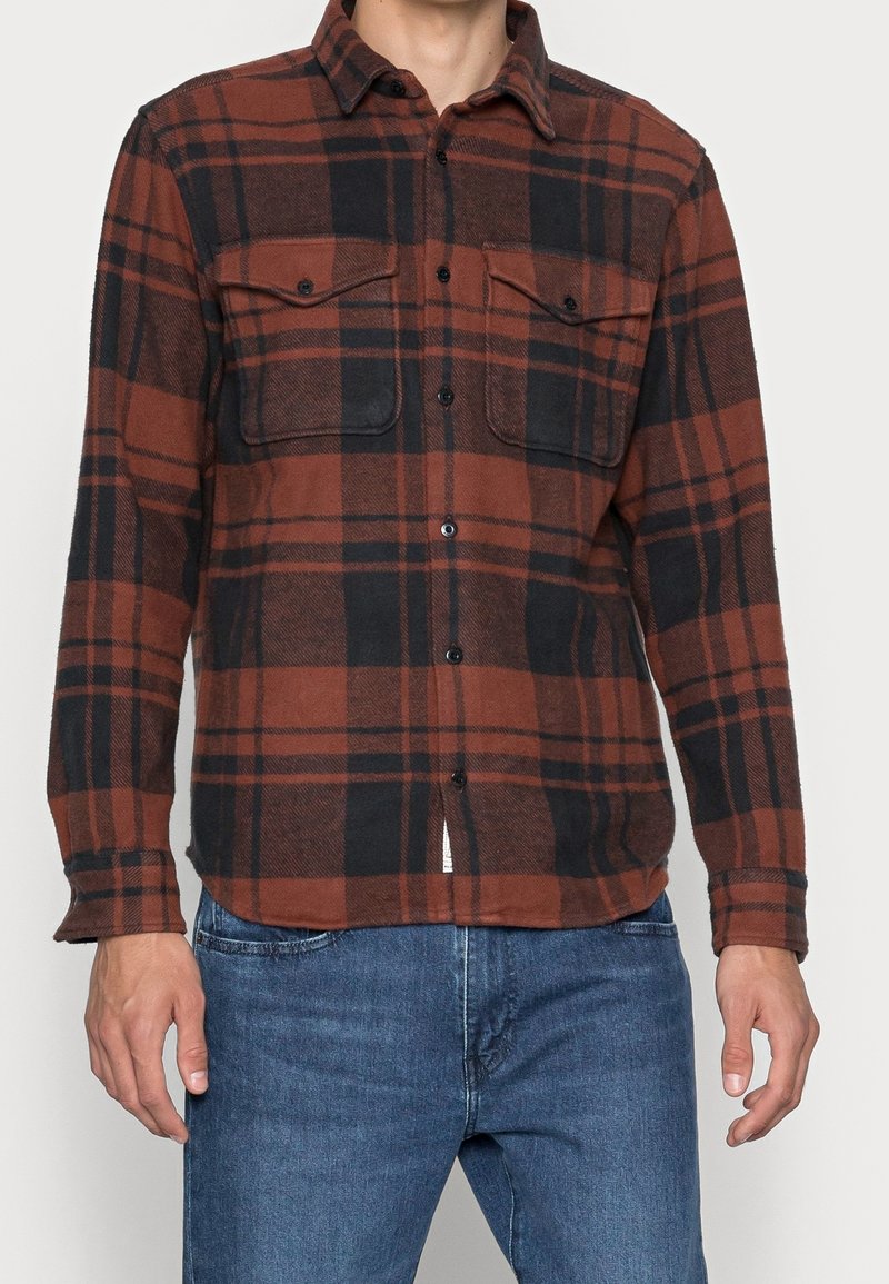 Flanelová košile v hnědém a černém kostkovaném vzoru, s dvěma kapsami na hrudi, zapínáním na knoflíky a dlouhými rukávy s manžetami na knoflíky. Texturovaná tkanina.