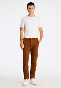 Pantalon en velours côtelé brun avec des côtes verticales, coupe droite, associé à un t-shirt blanc uni et des baskets blanches. Fond uni.