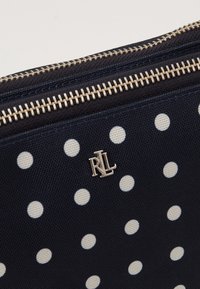 Marinblå canvaszippåse med vita prickar, med en silverfärgad logotypdetalj och guldglänsande dragkedja. Slät yta, kompakt design.