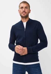 Marineblå zip-up cardigan med rund halsudsk�æring, ribbede manchetter og kant. Fremstillet af et blødt materiale, vist båret over en hvid skjorte.