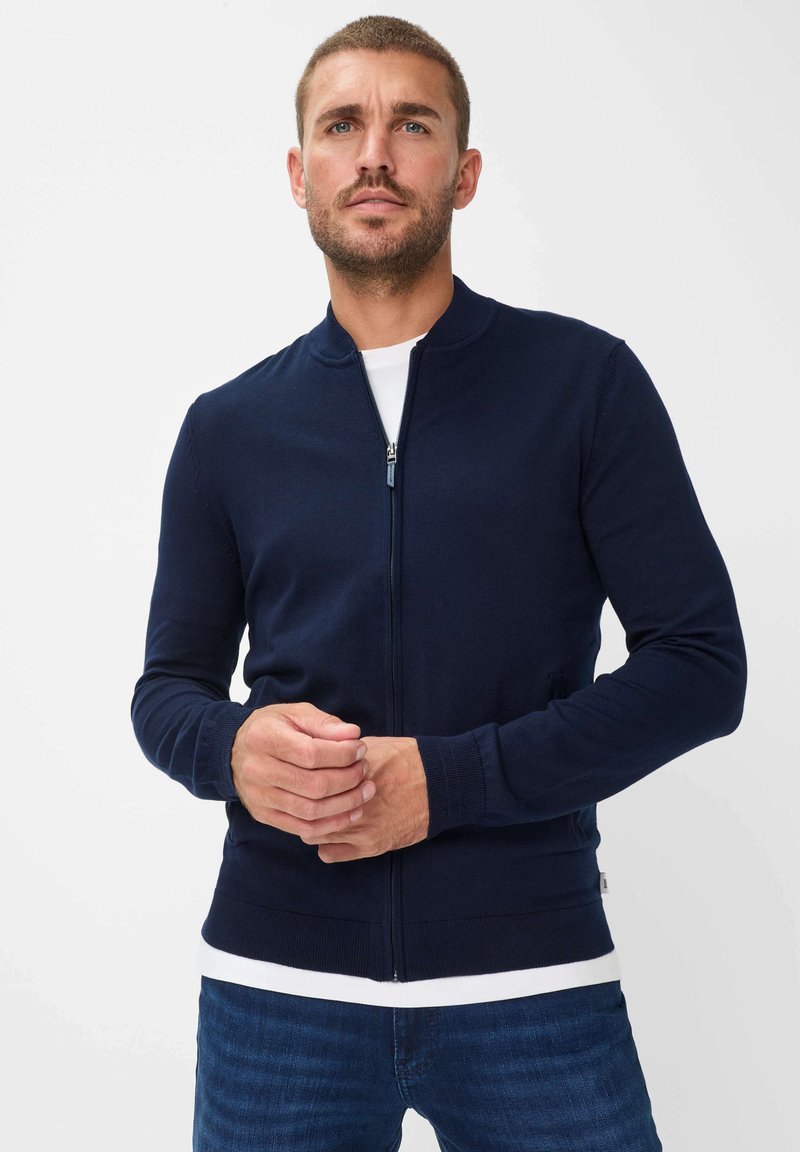 Marineblå zip-up cardigan med rund halsudskæring, ribbede manchetter og kant. Fremstillet af et blødt materiale, vist båret over en hvid skjorte.