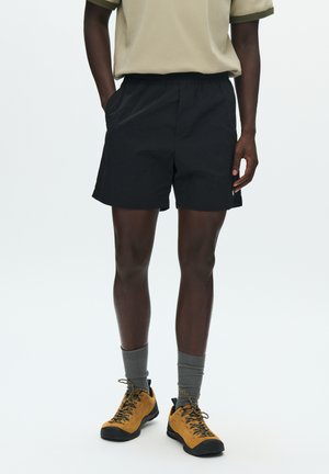 Pantaloncini neri con elastico in vita e tasche, realizzati in tessuto leggero. Abbinati a calzini grigi e scarpe da trekking color tan.