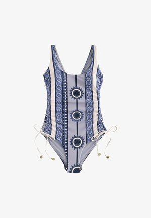 Femme aux longs cheveux blonds portant un maillot de bain bleu à motifs et un collier de coquillages, tenant un grand sac fourre-tout tissé, debout sur une plage rocheuse.