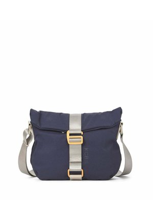 Persona que lleva una camiseta de talla suelta color beige y pantalones con zapatillas beige, llevando un bolso de hombro azul marino con correa beige y hebilla dorada.