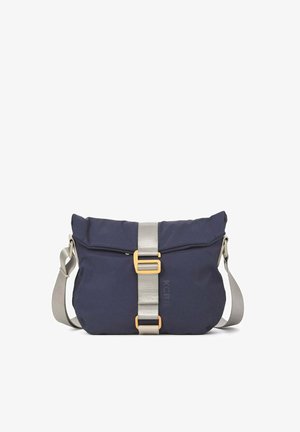 Personne portant un t-shirt ample beige et un pantalon avec des baskets beiges, portant un sac à bandoulière bleu marine avec une sangle beige et une boucle dorée.