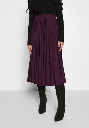 Woman wearing a long pleated dark purple skirt, black ribbed long-sleeve top, and black mid-calf boots, standing against a plain background.