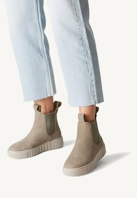 Bottines en suede kaki avec des panneaux latéraux élastiques et une semelle en caoutchouc striée, associées à un jean cropped bleu clair avec des ourlets effilochés.