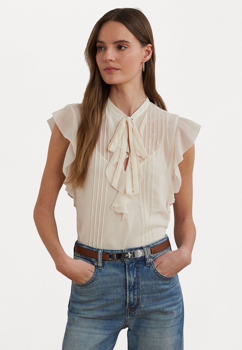 Cream blouse with ruffled sleeves, vertical pleats, and a tie at the neck. Worn with light blue denim jeans and a brown belt.