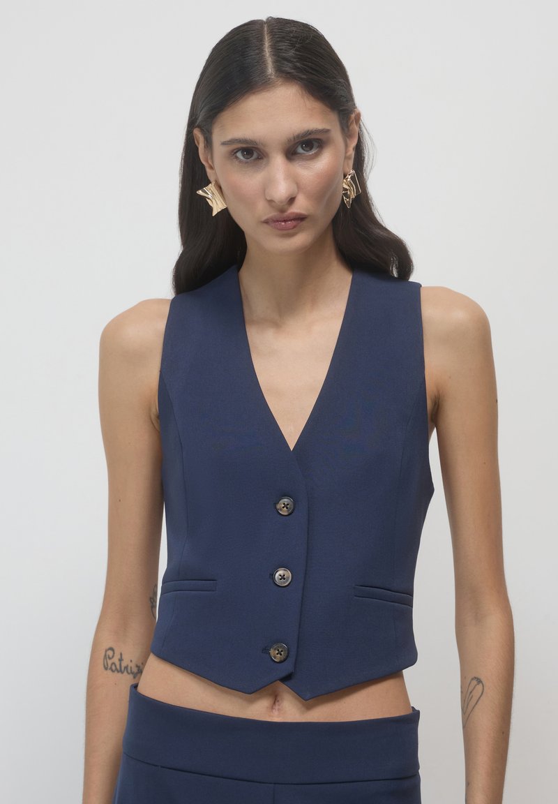 Marineblaue ärmellose Weste mit V-Ausschnitt, fünf Knöpfen und zwei Vordertaschen. Glatte Stoffqualität mit tailliertem Schnitt und cropped Saum.