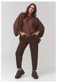 Brązowa koszula z guzikiem i obszernymi rękawami, w komplecie z dopasowanymi spodnimi spodniami i masywnymi czarnymi butami na sznurówki. Gładka faktura materiału.