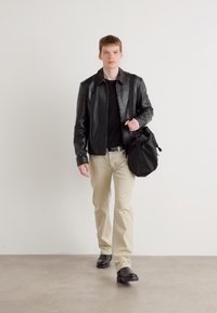 Chaqueta de cuero negra sobre una camisa negra, pantalones beige y zapatos negros. Lleva una bolsa negra. De pie contra un fondo simple.