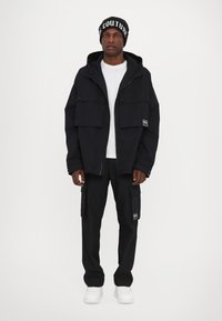 Black hooded jacket with large front pockets, worn over a white shirt and black cargo pants. Features a logo patch, paired with white sneakers.