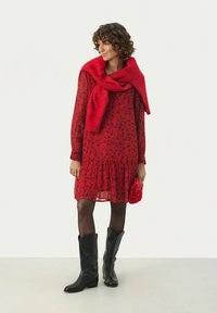 Rotes Blumenkleid mit einem Rüschensaum, transparenter Stoff, langen Ärmeln und einem gestrickten roten Pullover, der über die Schultern gelegt ist. Schwarze kniefreie Stiefel.