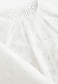 Blouse en dentelle blanche avec des motifs floraux, texture transparente et col rond avec une ouverture en arrière, fabriquée en tissu léger.