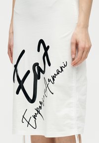 Λευκή φούστα με μεγάλο μαύρο λογότυπο "EA7 Emporio Armani" τυπωμένο διαγώνια, φορεμένη από άτομο ορατό από τη μέση έως το γόνατο.