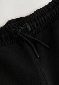 Černý elastický pás s tažným šňůrkou a plastovými zakončeními na černých látkových kalhotách.