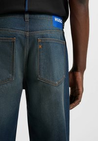 Jeans in denim blu scuro con cuciture arancioni e una toppa in pelle blu con marchio sulla cintura, dotati di due tasche posteriori con alette inclinate.