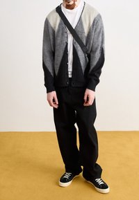 Knappkort cardigan i grå, svart och grädde med en diamantmönster, kombinerad med svarta byxor och svarta sneakers på ett gult golv.