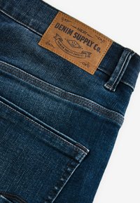Tamno plave traperice s kožnom etiketom na struku koja glasi "DENIM SUPPLY Co.", sa vidljivim šavovima i klasičnim dizajnom.