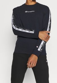 Sudadera de marina con cuello redondo, que presenta el logo "Champion" en blanco, acentos de rayas negras y blancas en las mangas con texto de logo, y puños acanalados.