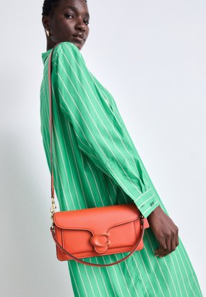 Femme portant une robe rayée verte avec un petit sac à bandoulière en cuir orange avec des pièces métalliques dorées sur un fond clair uni.