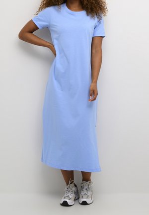 Mujer que lleva un vestido midi de manga corta azul claro y zapatillas deportivas blancas, de pie con una mano en la cadera con un fondo liso.