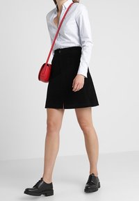 Gonna di velluto a coste nera con uno spacco frontale, abbinata a una camicia botton-down a fantasia azzurro chiaro e scarpe stringate nere, mano che tiene una borsa a tracolla rossa.