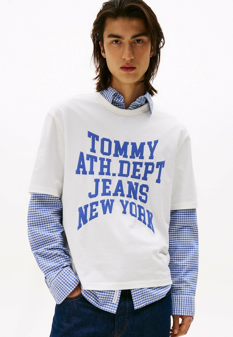 Hvid bomulds T-shirt med blå grafisk tekst, der læser "TOMMY ATH. DEPT JEANS NEW YORK," lagdelt over en blå og hvid ternet skjorte.