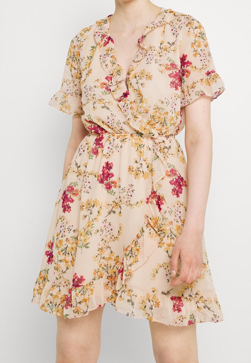 Kukkakuvioinen kietaisumekko pehmeässä beigemäisessä sävyssä, jossa on röyhelömuotoiset hihat ja helma, koristeltuna vaaleanpunaisilla ja keltaisilla kukilla, valmistettu kevyestä kankaasta.