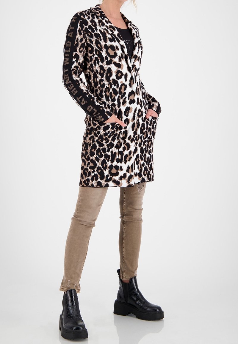 Femme portant un long blazer imprimé léopard avec le texte « WILD AT HEART » sur les manches, un pantalon beige slim et des bottines noires à plateforme.