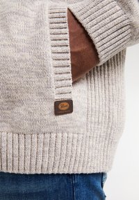 Béžový pletený svetr s ribovaným lemem a manžetami. Obsahuje hnědou koženou nášivku s logem. Pod ním je částečně viditelná hnědá košile.