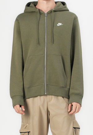 Personne portant un sweat à capuche zippé Nike vert olive avec capuche et pantalon cargo beige, debout les bras détendus le long du corps.