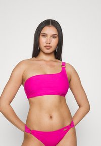 Top de biquíni rosa com um ombro só, com uma textura suave e um detalhe em ouro na alça. As partes de baixo combinando apresentam um laço lateral.