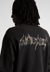 Hajlakkos férfi fekete pólót visel, amelyen sárga és lila stilizált, cakkos szövegtervezés található a hátán.