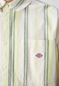 Camisa de manga corta a rayas en beige, amarillo y verde. Presenta un cuello con botones, bolsillo en el pecho y una etiqueta de "Dickies". Material de algodón.