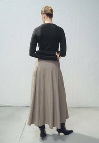 Top negro de manga larga combinado con una falda plisada beige. Lleva botas negras de tacón alto. Texturas suaves y colores neutros. Vista desde atrás.