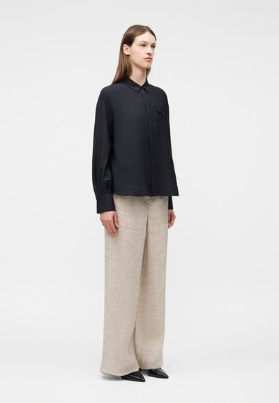 Γυναίκα που στέκεται φορώντας μια μαύρη φαρδιά μακρυμάνικη μπλούζα, μπεζ παντελόνι με φαρδιά μπατζάκια και μαύρα μυτερά παπούτσια σε απλό φωτεινό φόντο.