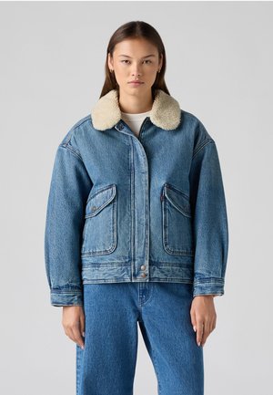 Denim jaka ar bežīgu mākslīgo kažokādu apkakli, priekšējiem kabatām un brīvu piegriezumu. Gaiši zils tonis ar redzamām šuvju detaļām un metāla pogām.