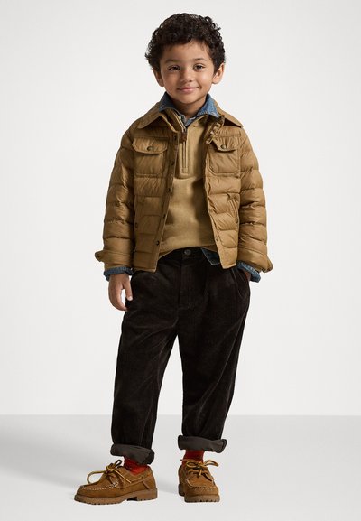 Polstret brun jakke med trykknapp-lommer, beige genser, mørke kordfløyel bukser og brune snøresko. Barnet smiler mot en ensfarget bakgrunn.