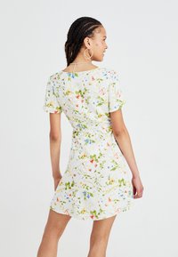 Bloemjurk met korte mouwen, witte achtergrond. Heeft een kleurrijke botanische print, een aansluitende silhouet en een subtiele opening aan de achterkant met beslag.
