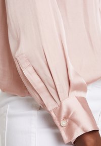 Ljusrosa satinblus med långa ärmar, knappade manschetter och en slät textur. Skuren med en lätt drapering för en elegant passform.