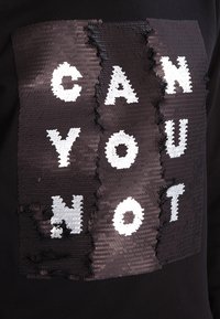 Sudadera negra con un diseño de lentejuelas que presenta el texto "NO PUEDES" en blanco y plateado. El texto está resaltado con texturas contrastantes.