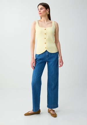 Femme debout, portant un haut sans manches jaune pâle boutonné, un jean bleu, des ballerines marron et des boucles d'oreilles dorées, regardant sur le côté.